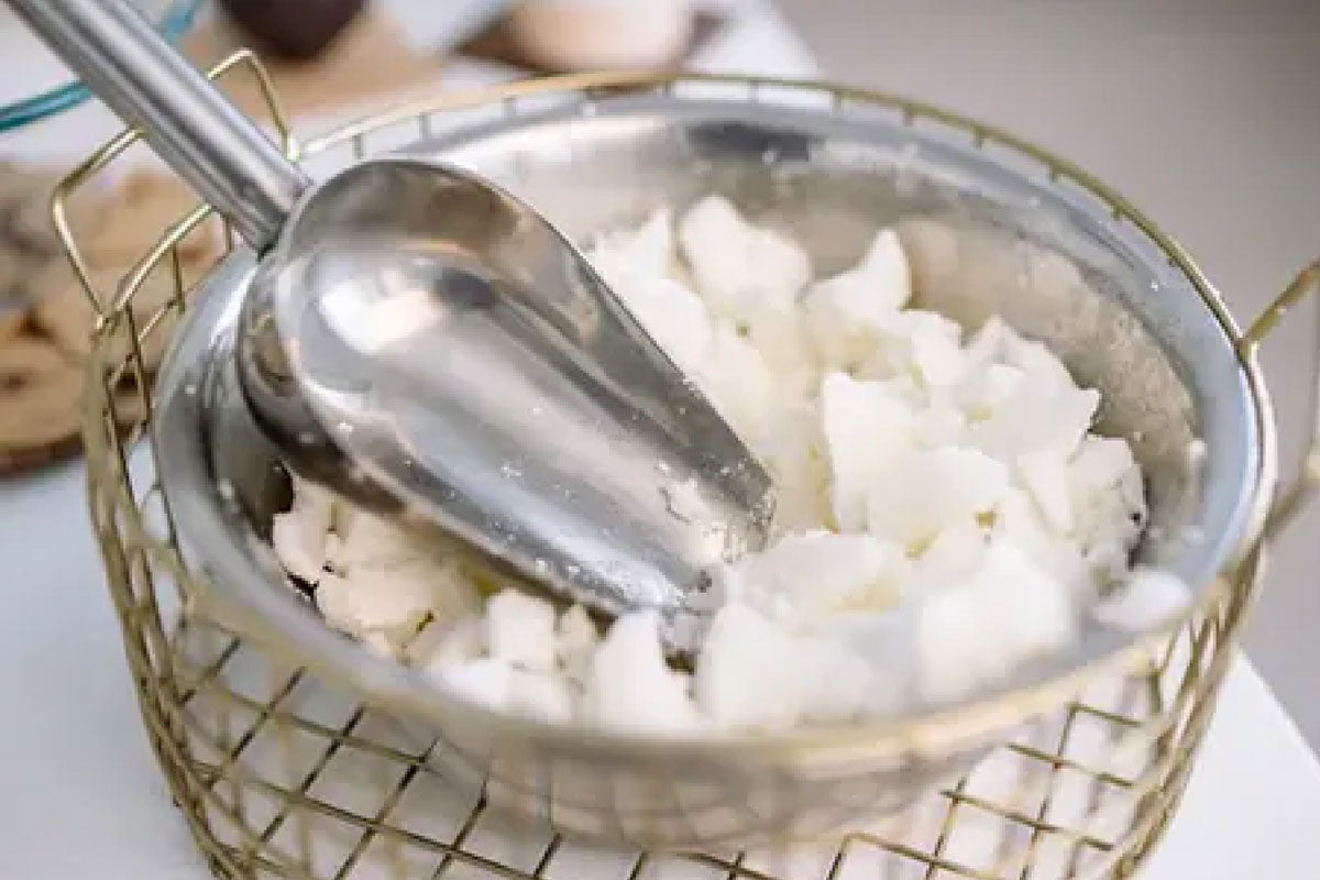 A metal scoop resting inside a stainless steel bowl filled with chunks of un-melted white candle wax. The bowl sits inside a gold wire basket on a white surface.
