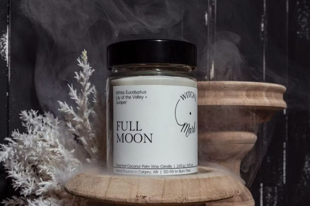 A Witch's Mark 'Full Moon' candle featuring notes of white eucalyptus, lily of the valley, and juniper, resting on a carved wooden pedestal surrounded by white dried botanicals and atmospheric smoke.
