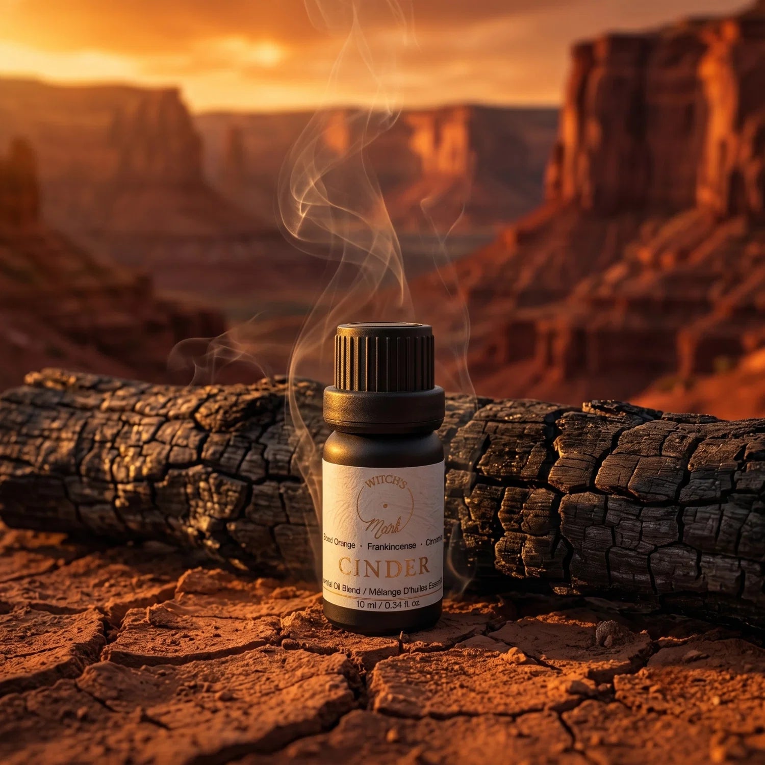Bottle of 'Cinder' essential oil blend next to a charred log with a desert landscape at sunset.