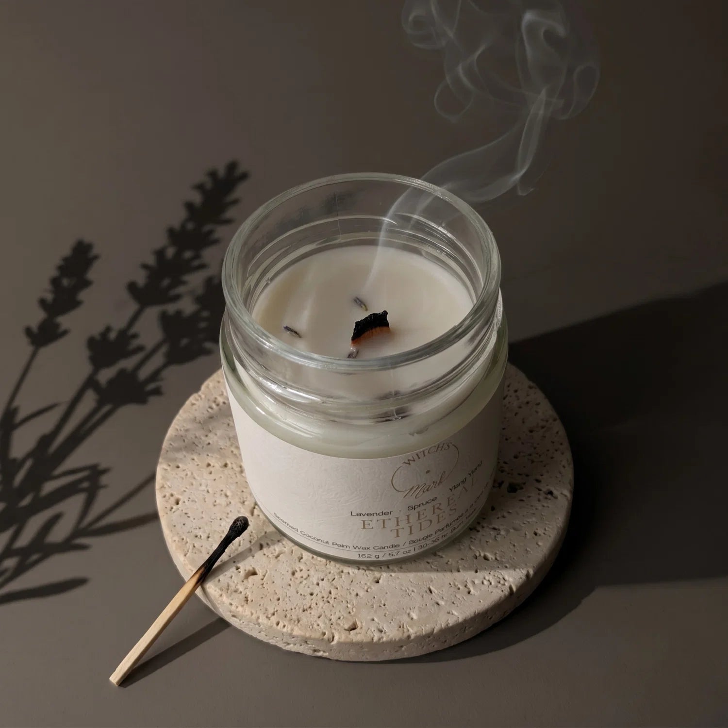 Ethereal Tides candle recently extinguished with whirls of smoke coming from the wick. In the background is a shadow of a sprig of lavender.
