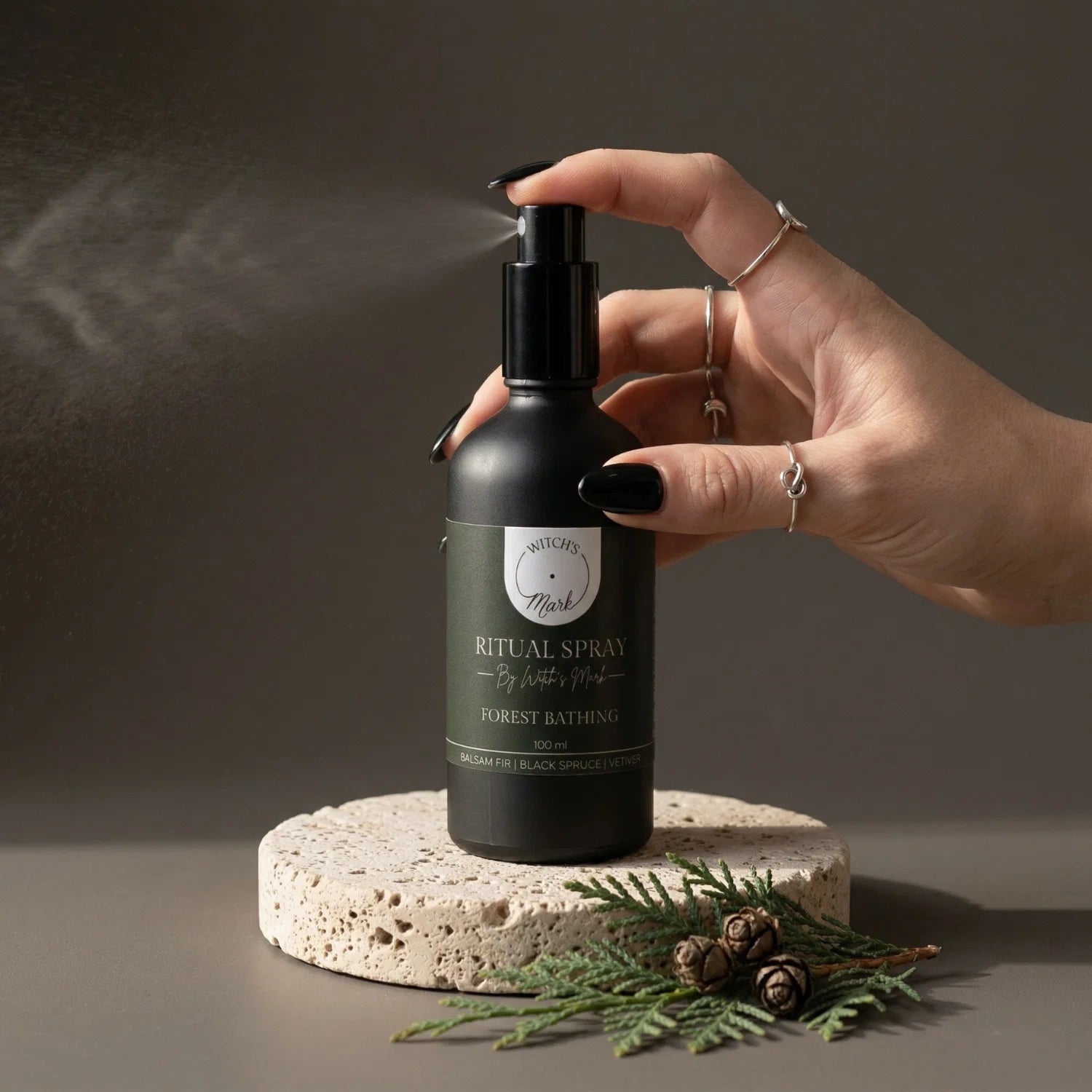 A hand with black nail polish spritzing the Witch's Mark Forest Bathing ritual spray over fresh evergreen sprigs and tiny pinecones. This grounding aura mist is infused with tree agate charged moon water and woody essential oils to anchor your wild spirit and center the mind.