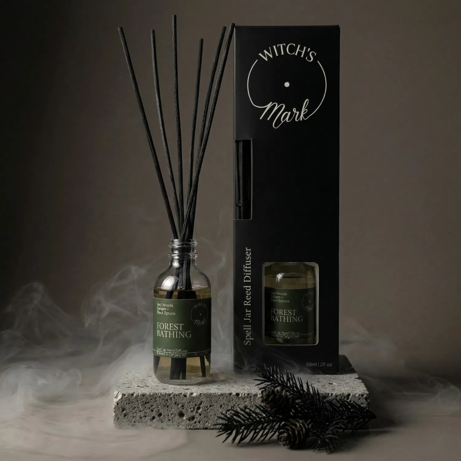 The Forest Bathing spell jar reed diffuser by Witch's Mark displayed next to its sleek black packaging box. The apothecary glass vessel with black reeds rests on a textured stone block surrounded by mystical mist and a fresh black spruce sprig, crafted to deeply root your energy.
