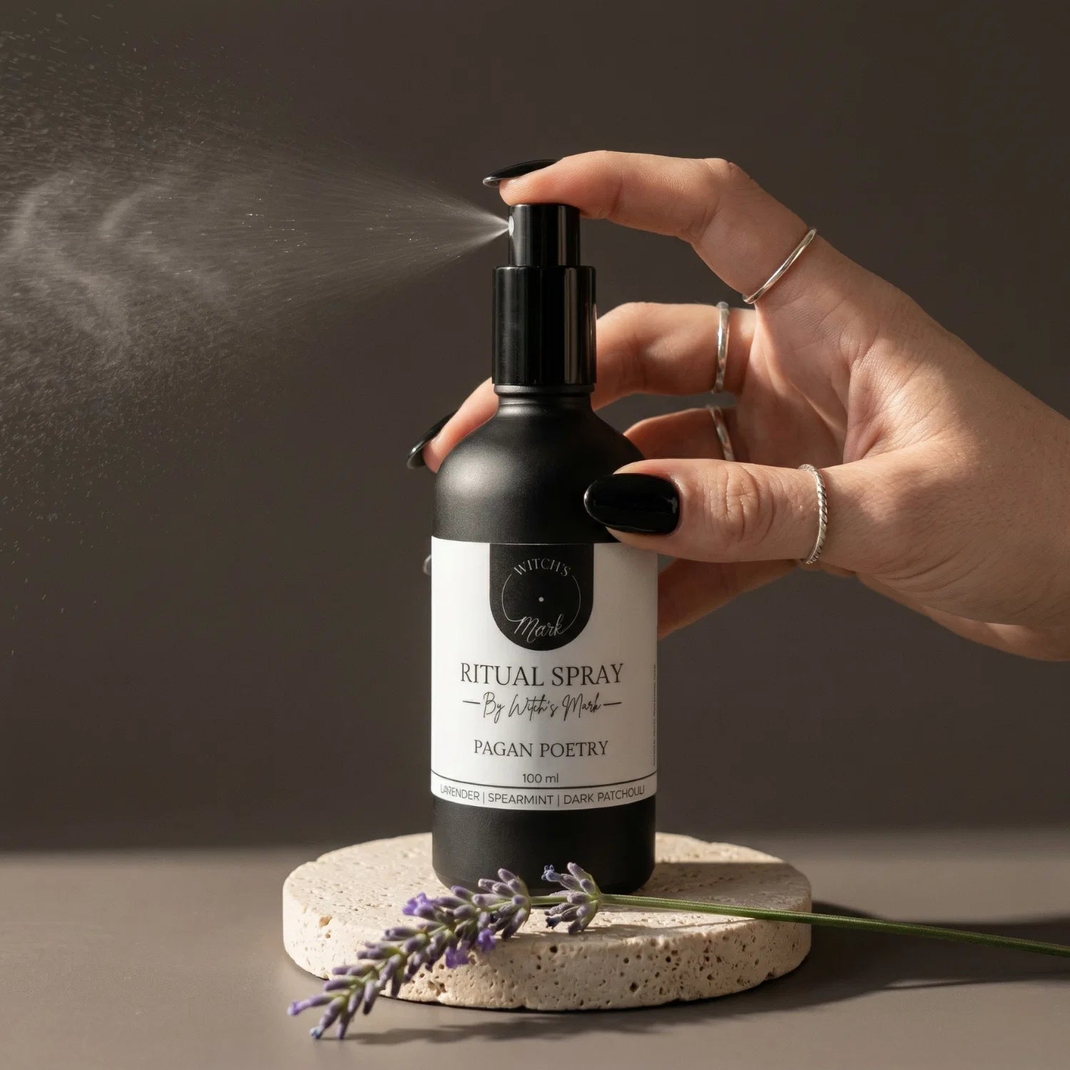 A hand with black nail polish spritzing the Witch's Mark Pagan Poetry ritual spray over a fresh sprig of purple lavender. This calming aura mist is infused with amethyst moon water and essential oils to quiet the mind and invoke absolute tranquility.