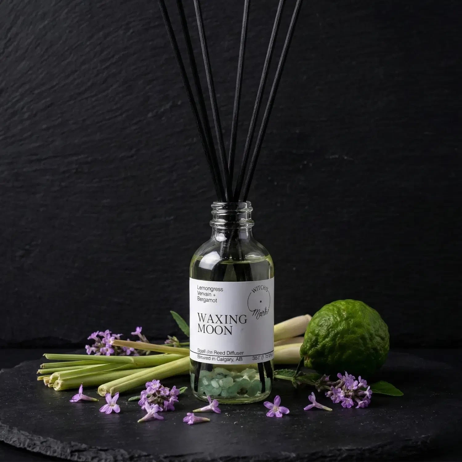 The Waxing Moon spell jar reed diffuser by Witch's Mark showcased against a dark slate background, surrounded by its core botanical ingredients: fresh lemongrass stalks, a whole green bergamot citrus, and delicate purple vervain flowers.