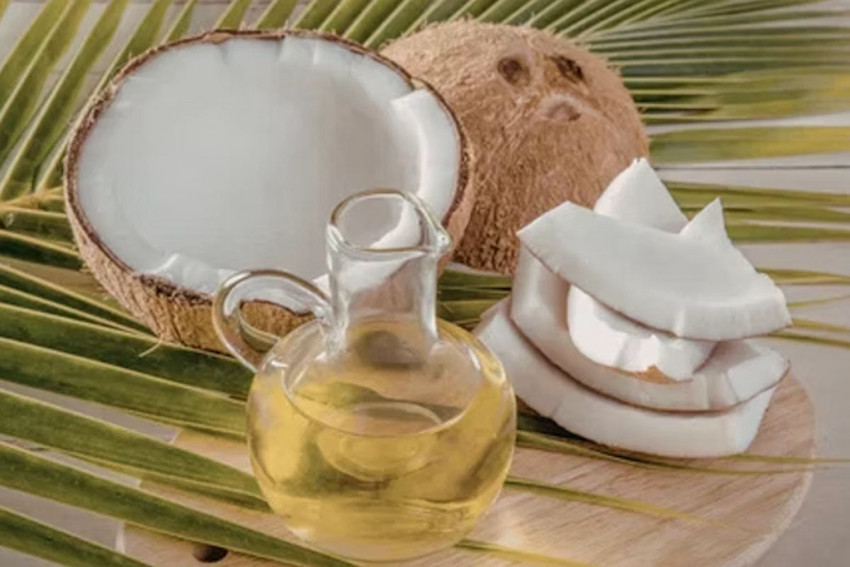 A small glass pitcher of clear coconut oil sitting next to fresh coconut halves and sliced white coconut meat on a green palm leaf.