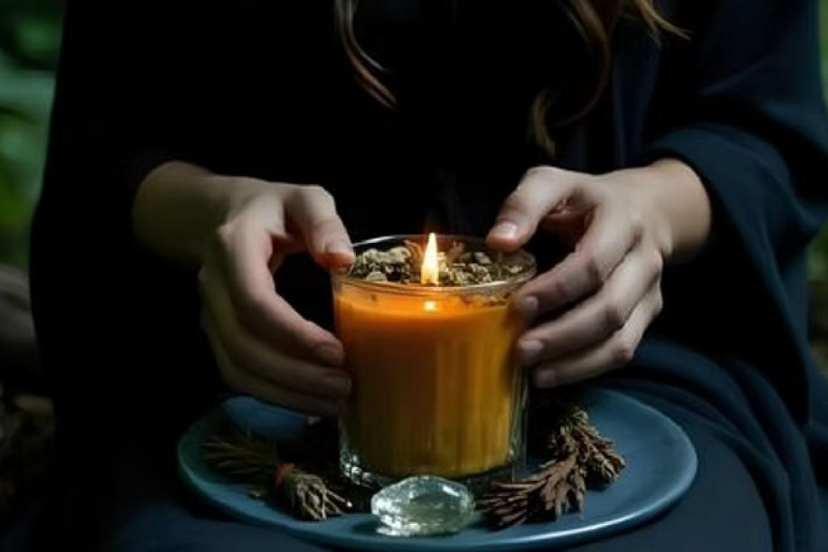 A person's hands gently framing a glowing, lit orange candle in a glass jar. The candle is topped with dried herbs and sits on a dark plate next to small bundles of dried cedar and a clear quartz crystal.