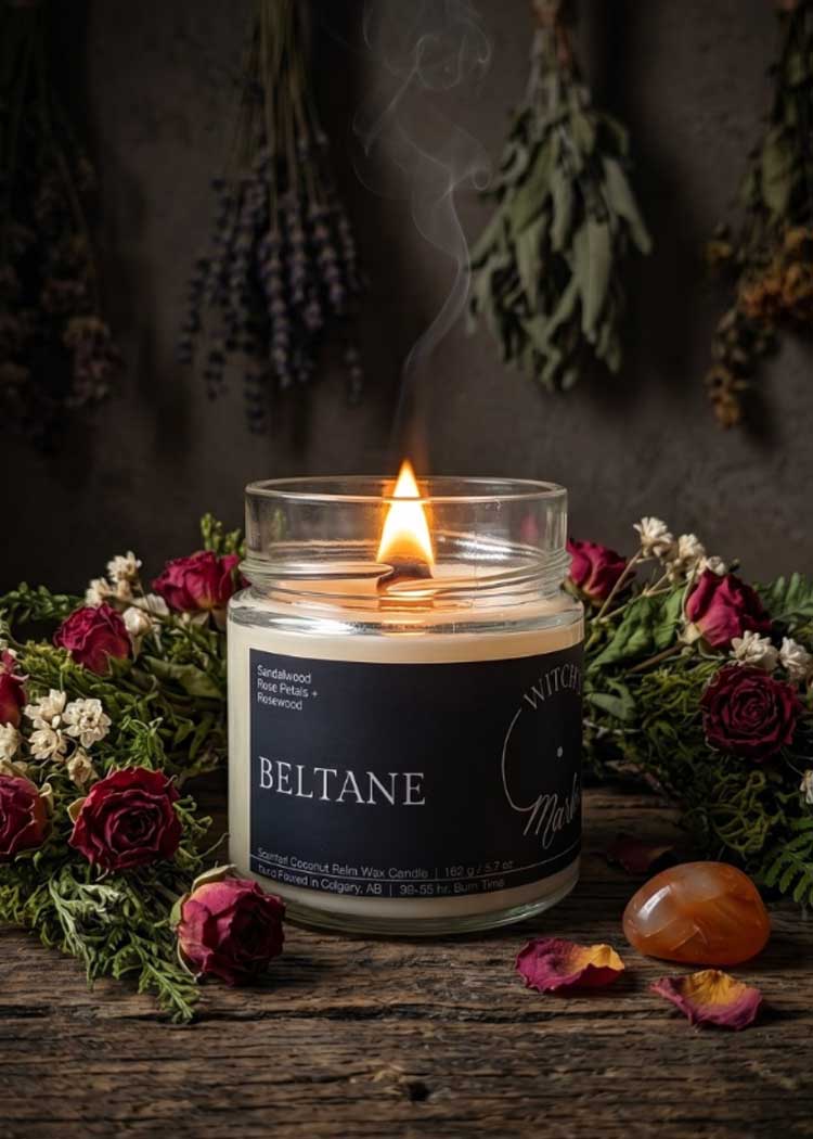 A lit Witch's Mark Beltane ritual candle on a rustic wooden surface, surrounded by dried red roses, botanical elements, and a carnelian crystal stone.
