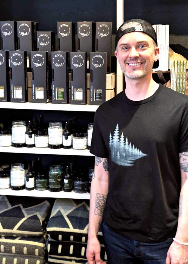 A smiling portrait of Daryl, the witch and founder of Witch's Mark, standing in front of shelves stocked with handmade eco-friendly home fragrance products at a local market.