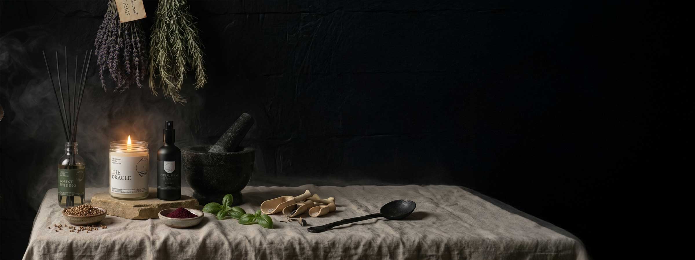 A panoramic, atmospheric display for the Witch's Mark Essential Rituals collection, featuring a lit 'The Oracle' intention candle, a 'Forest Bathing' reed diffuser, and a ritual spray on a linen tablecloth alongside a stone mortar and pestle, dried hanging herbs, and loose ritual ingredients.