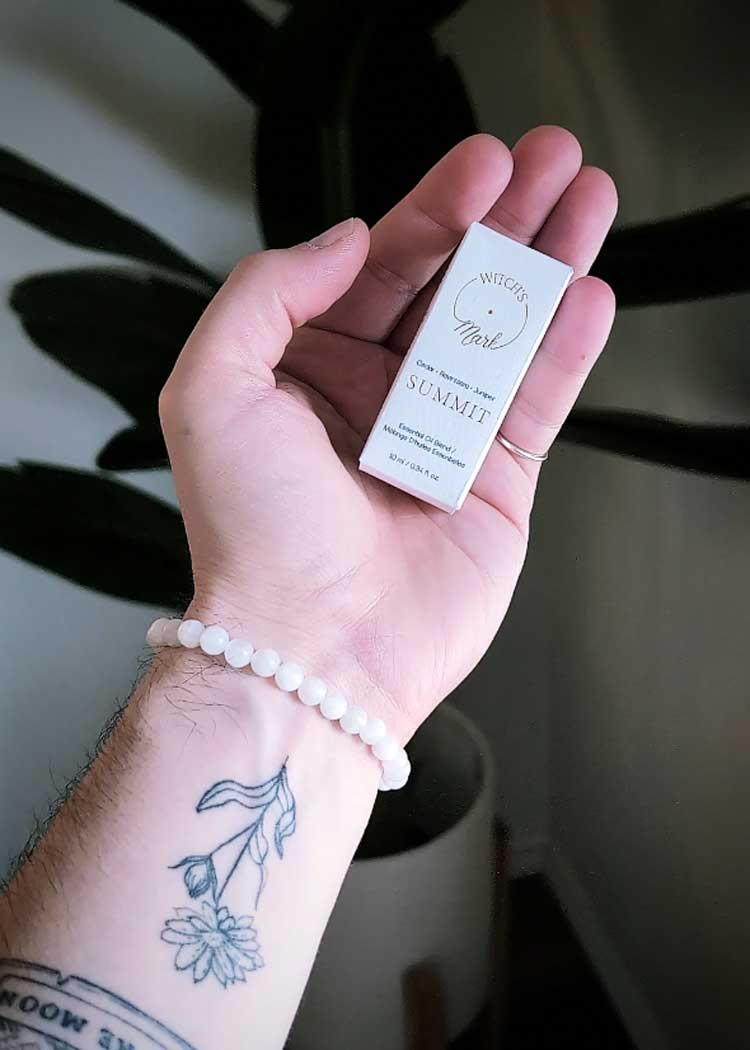 A close-up of a hand with a botanical wrist tattoo and crystal bracelet holding a small white Witch's Mark product box against a soft background.
