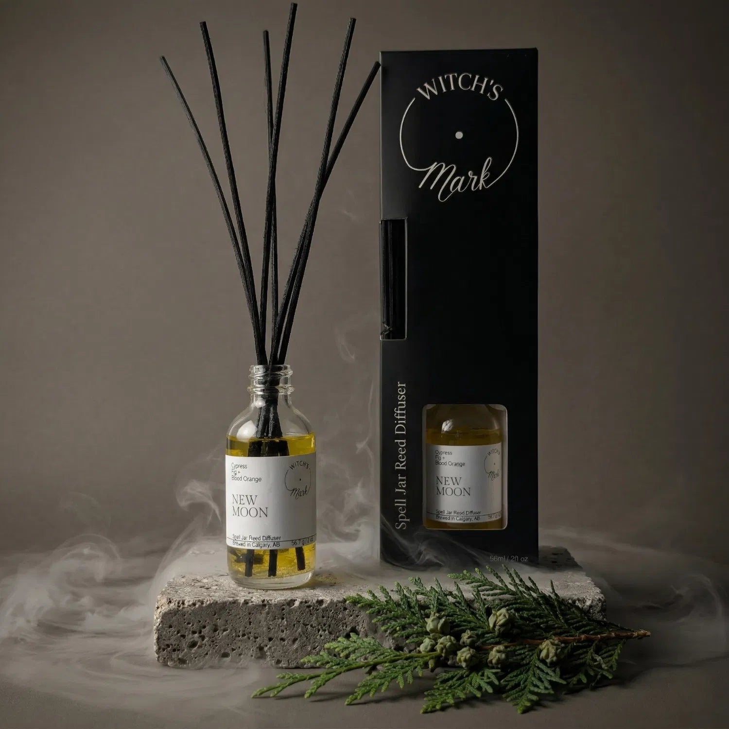 The New Moon spell jar reed diffuser by Witch's Mark displayed alongside its sleek black packaging box. The glass apothecary vessel with black reeds rests on a textured stone block amidst a mystical mist, garnished with a fresh grounding cypress sprig.