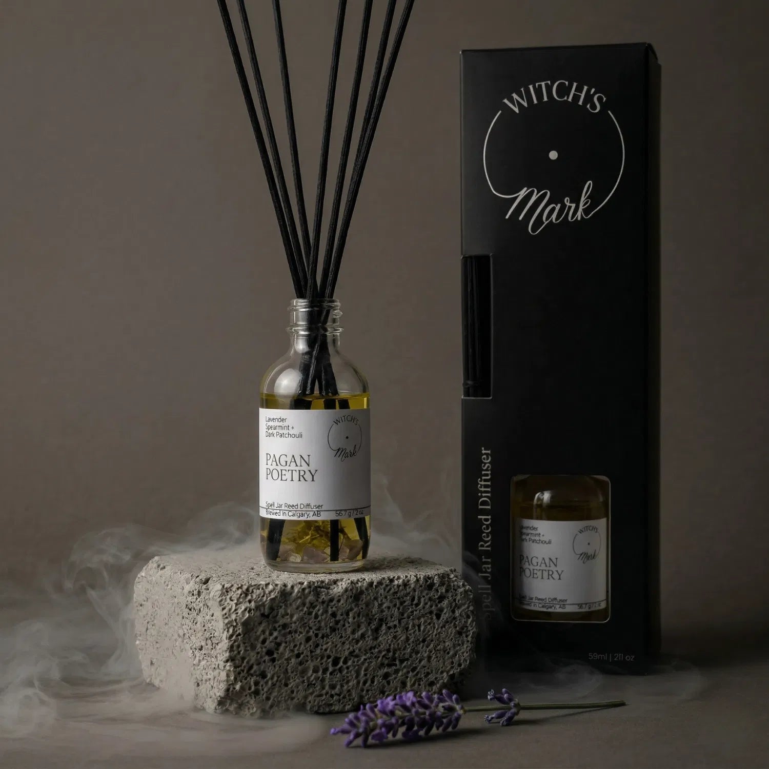 The Pagan Poetry spell jar reed diffuser by Witch's Mark displayed next to its sleek black packaging box. The apothecary glass vessel with black reeds rests on a textured stone block surrounded by mystical mist and a fresh sprig of calming lavender.