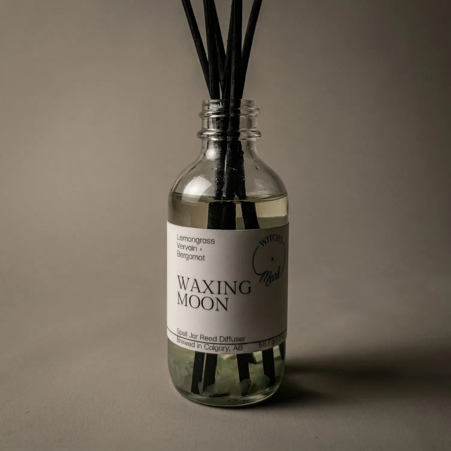 A dramatic spotlight close-up of the Waxing Moon spell jar reed diffuser, highlighting the front label which lists its magical, momentum-building scent profile of lemongrass, protective vervain, and bergamot.