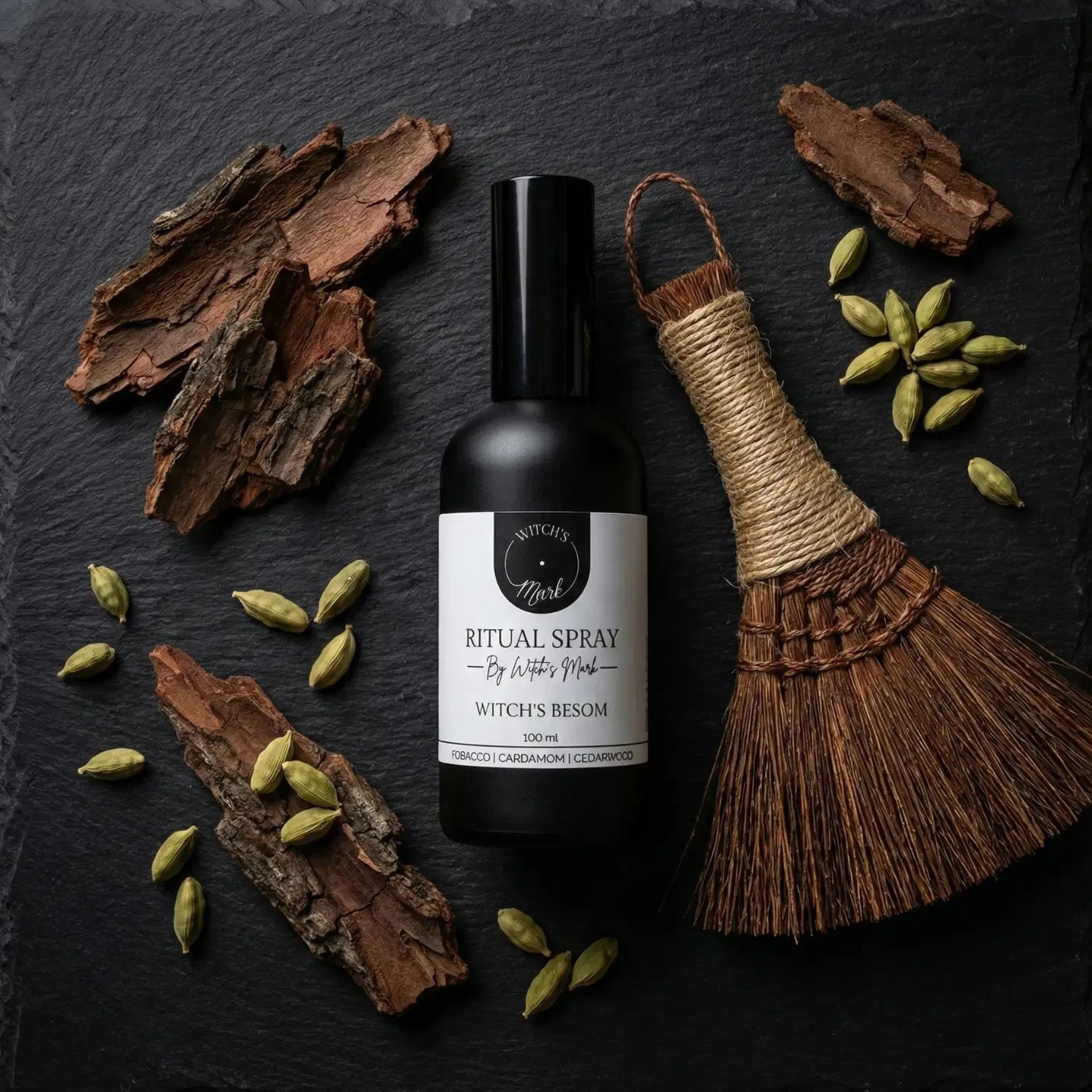 A top-down flat lay highlighting the space-clearing ingredients of the Witch's Besom ritual mist by Witch's Mark. The black apothecary bottle sits surrounded by a miniature sweeping besom, pieces of deep resinous cedarwood, and scattered green cardamom pods against a dark slate background.